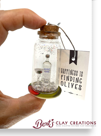 Hand holding a small glass jar with a figurine inside, featuring a tag with text and 'Bert's Clay Creations' branding.